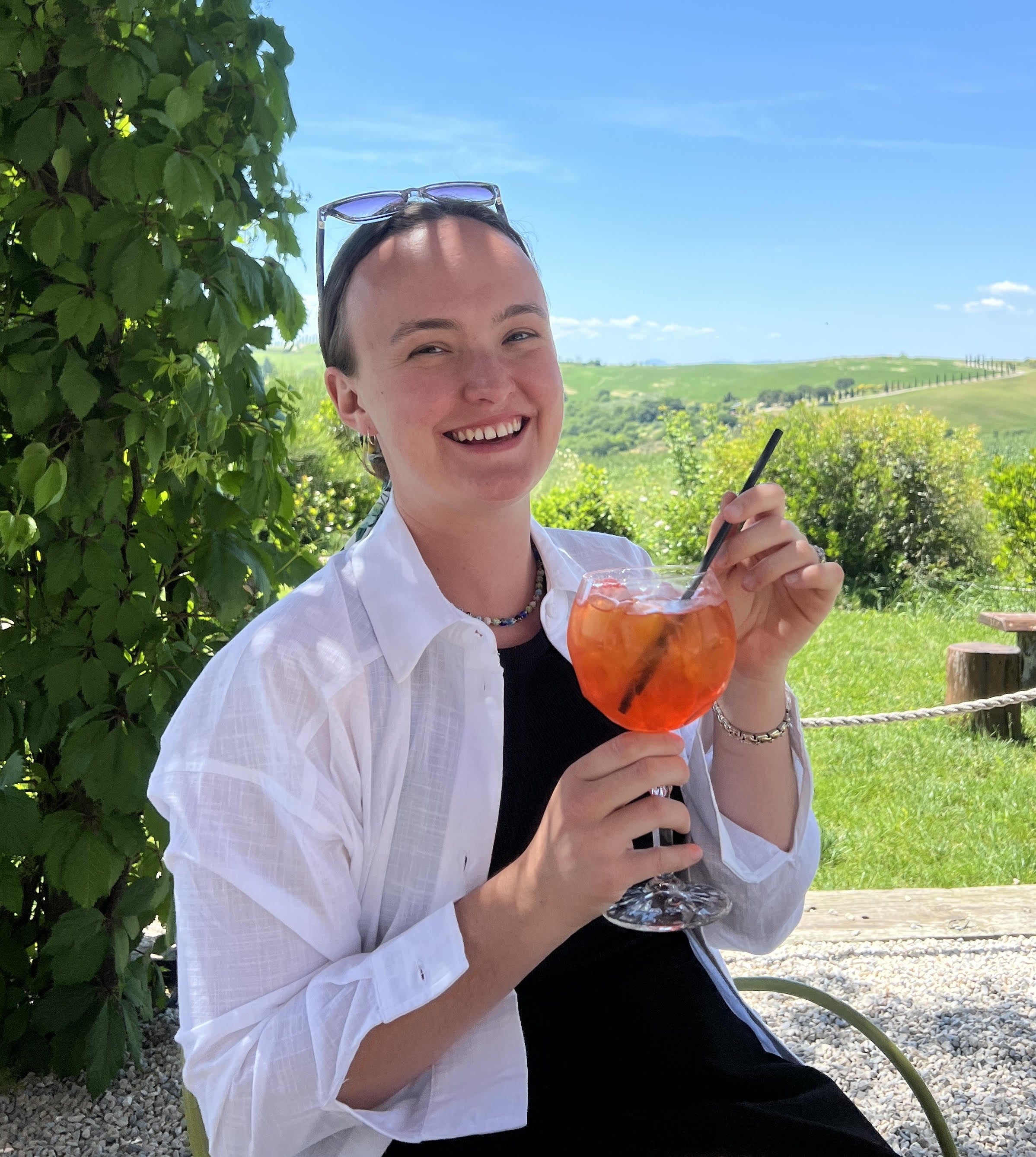 Woman holding an orange drink called an Aperol spritz with a scenic background of Tuscany's Val d'Orcia region.