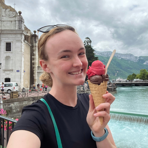 Woman holding a pink ice cream cone with chocolate topping in front of a scenic background with mountains and buildings. Location is Lake Annecy France. 