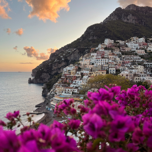Village perched on a cliff overlooking the ocean with purple flowers in the foreground. Location is Positano, Amalfi Coast, Italy.